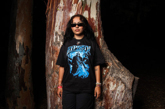 Female model wearing Hypnotic Clothing ‘The Scythe’ blue graphic streetwear tee standing in front of a tree at night.