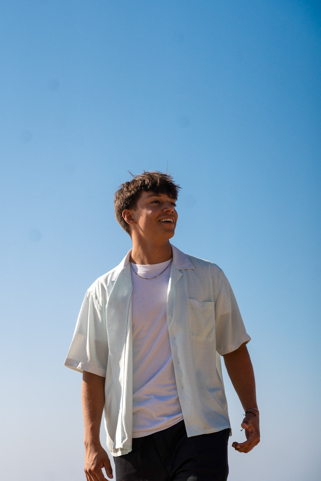 Male model wearing the Hypnotic Clothing HYP light blue button-up shirt standing against a bright coastal sky, modern casual streetwear.