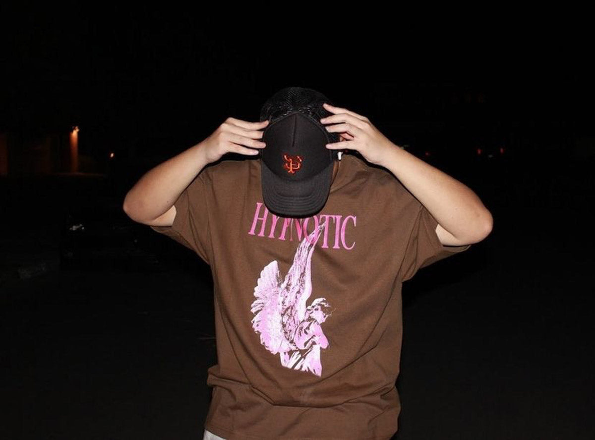 Model wearing Hypnotic Clothing ‘Angel’ brown graphic tee with pink angel artwork, posing outdoors at night.