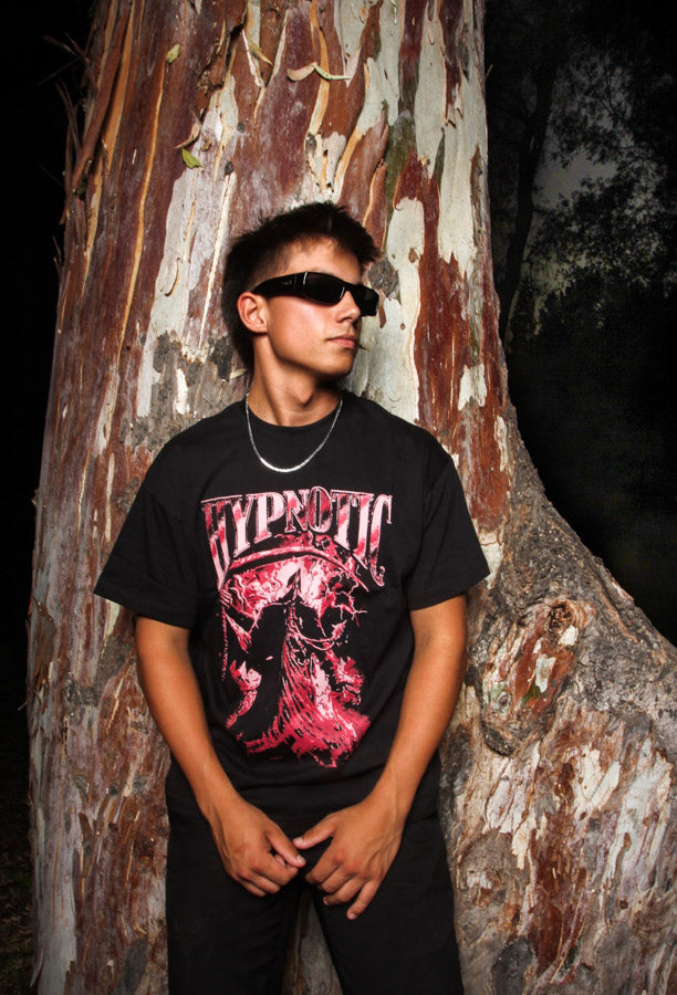Male model wearing Hypnotic Clothing ‘The Scythe’ red graphic streetwear tee while standing in front of a textured tree at night.