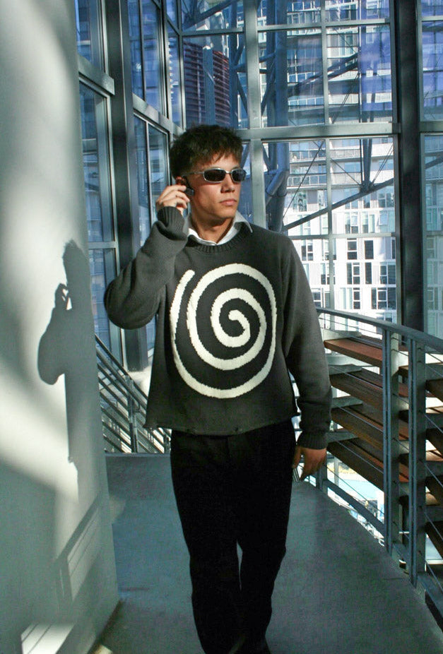 Full-body shot of model wearing the Hypnotic Clothing Lost in the Matrix spiral sweater in glass atrium, contemporary streetwear look.