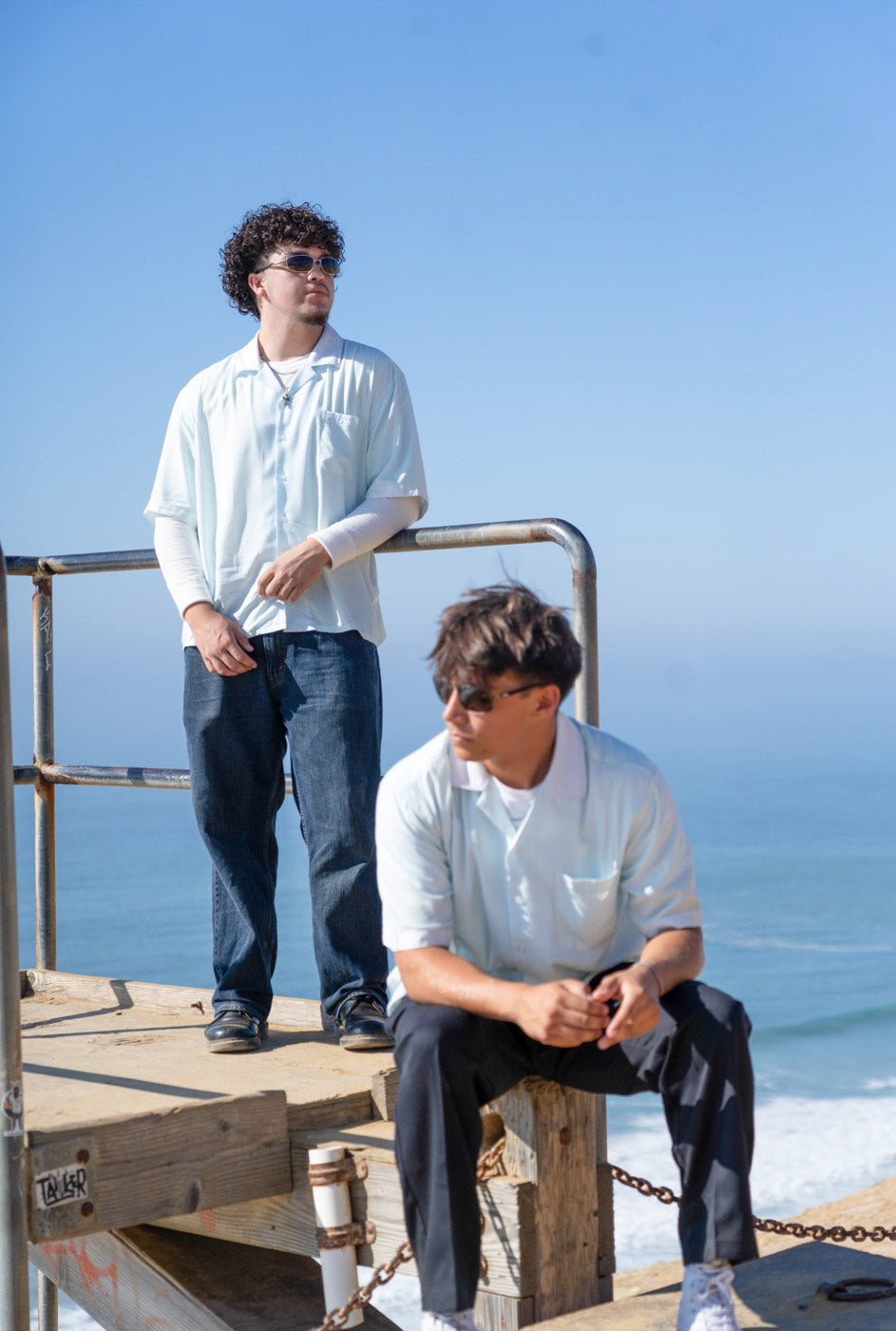 Two models wearing the Hypnotic Clothing HYP button-up shirt sitting and standing by a coastal railing, light minimal streetwear aesthetic.