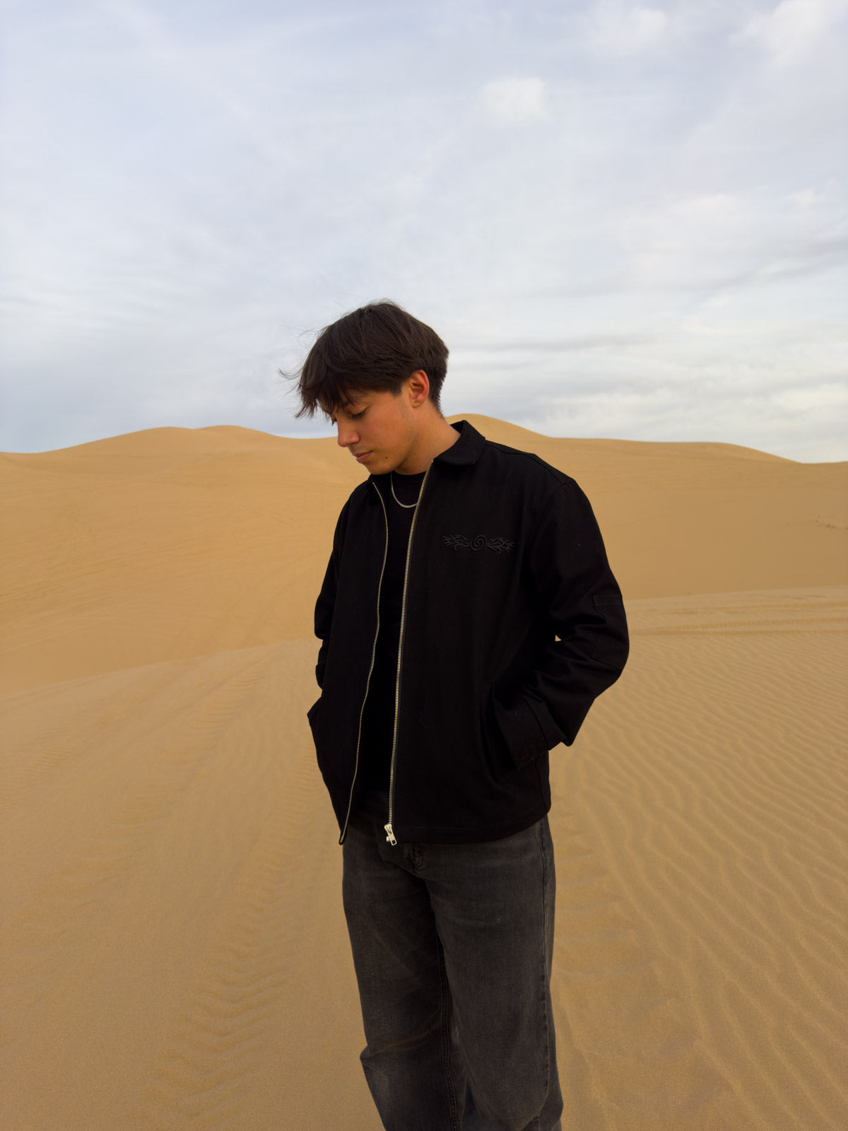 Male model wearing the Hypnotic Clothing Lucien black work jacket standing in desert sand dunes, modern streetwear portrait.