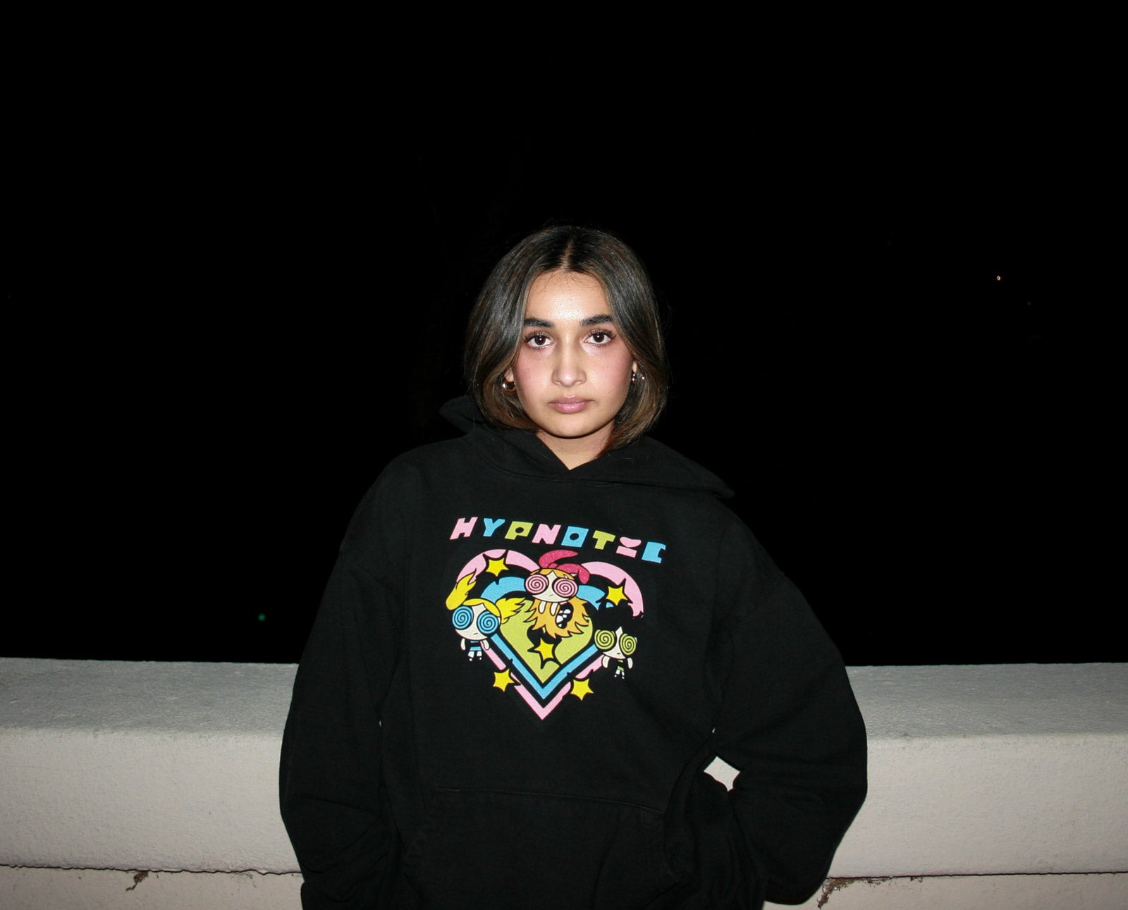 Female model wearing the Hypnotic Clothing PPG graphic hoodie in nighttime setting, vibrant nostalgic streetwear design.
