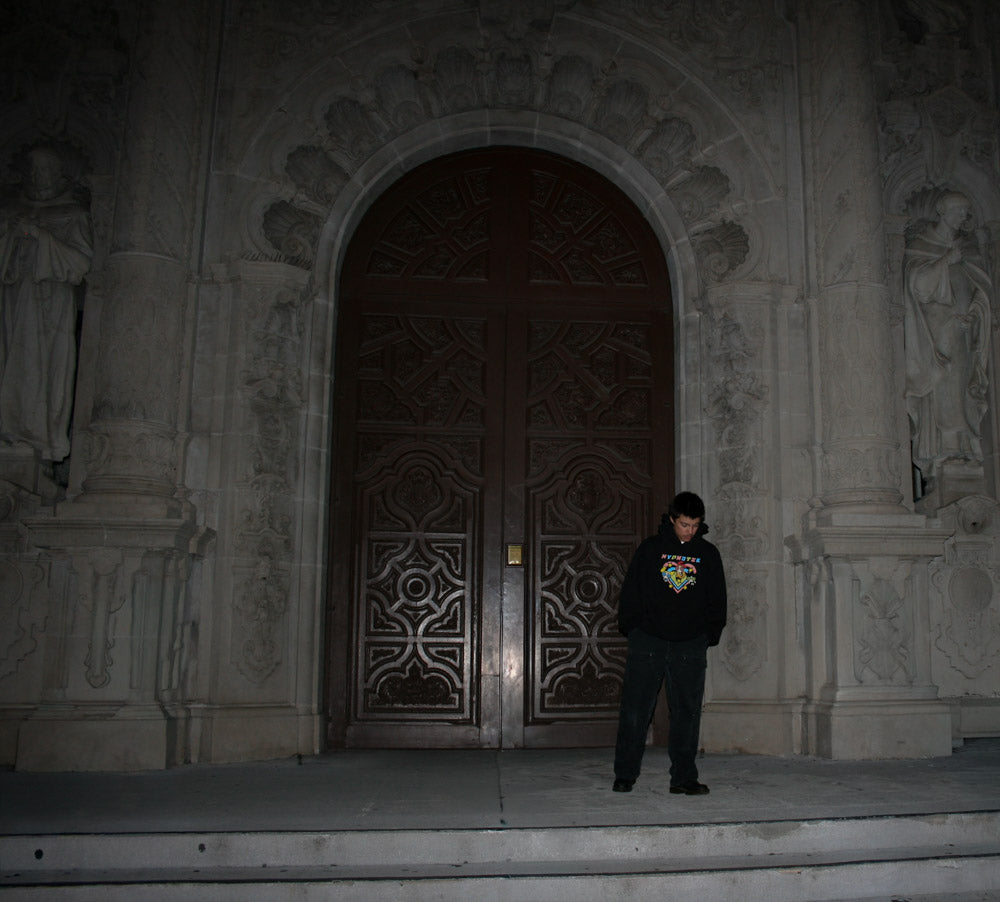 Model wearing the Hypnotic Clothing PPG graphic hoodie standing before ornate carved doors at night, cinematic streetwear photography.