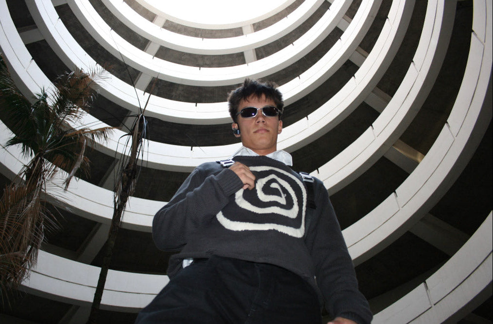 Male model wearing the Hypnotic Clothing Lost in the Matrix spiral sweater beneath circular parking garage architecture, dramatic streetwear shot.