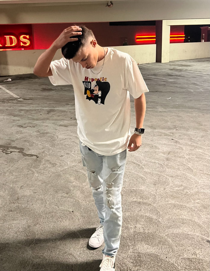Person wearing Hypnotic Clothing ‘Mickey’ white streetwear T-shirt standing in a lit parking garage at night.
